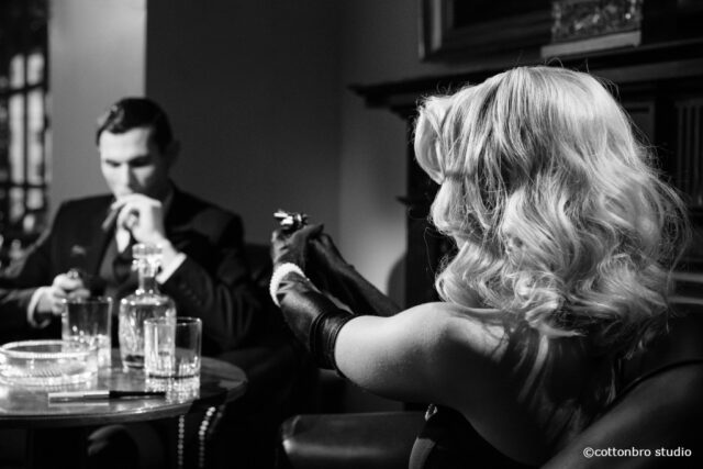 Schwarz-weiß-Bild einer Dame und eines Herren im Salon. Sie hält eine Waffe auf ihn gerichtet.
